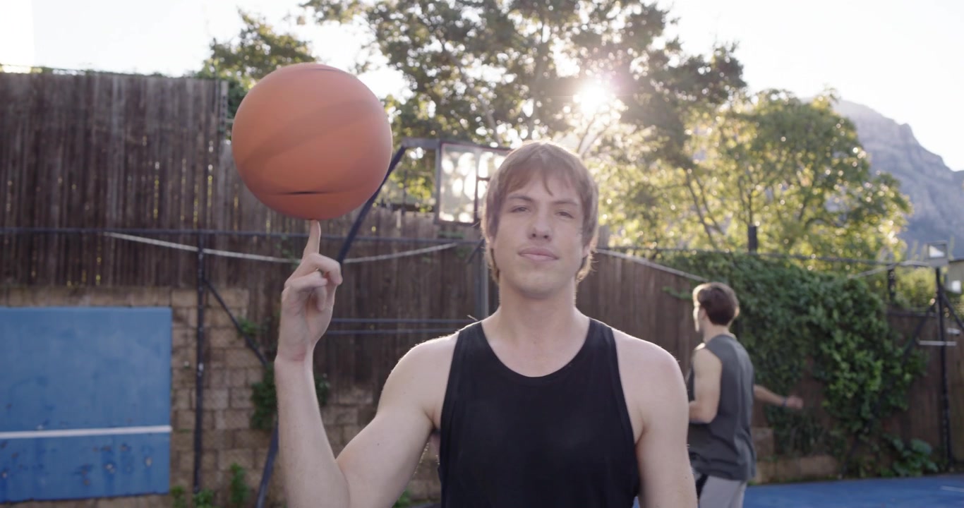 Male friends on backyard court, sunlit tall adult in tank top practicing spins on orange basketball