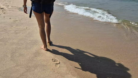 barefoot female backpacker hikes on beach sand near gentle waves