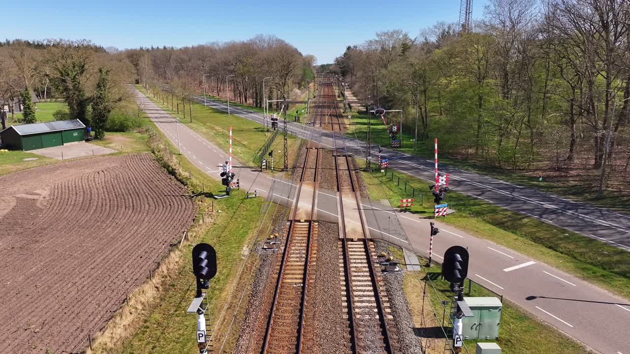 Static drone view shows barriers closing at a railway crossing on a countryside road, green fields, trees, and tracks visible, rural intersection in the Netherlands, real time, spring daylight.