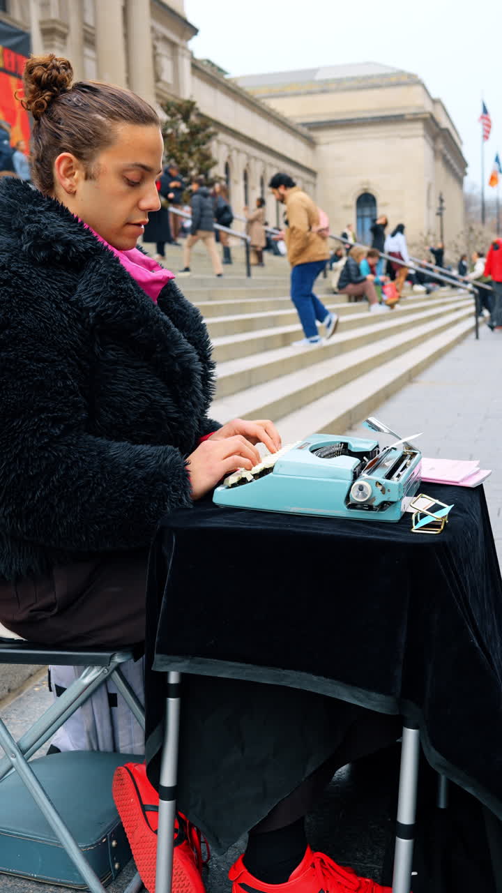 New York, USA, 2 Maret 2026: Tangan seorang penulis menggunakan mesin tik manual di luar ruangan. Pemandangan jarak dekat seseorang mengetik di mesin tik vintage biru yang duduk di tangga sebuah bangunan umum di New York
