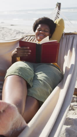 video vertical de una mujer afroamericana leyendo un libro en una hamaca en la playa