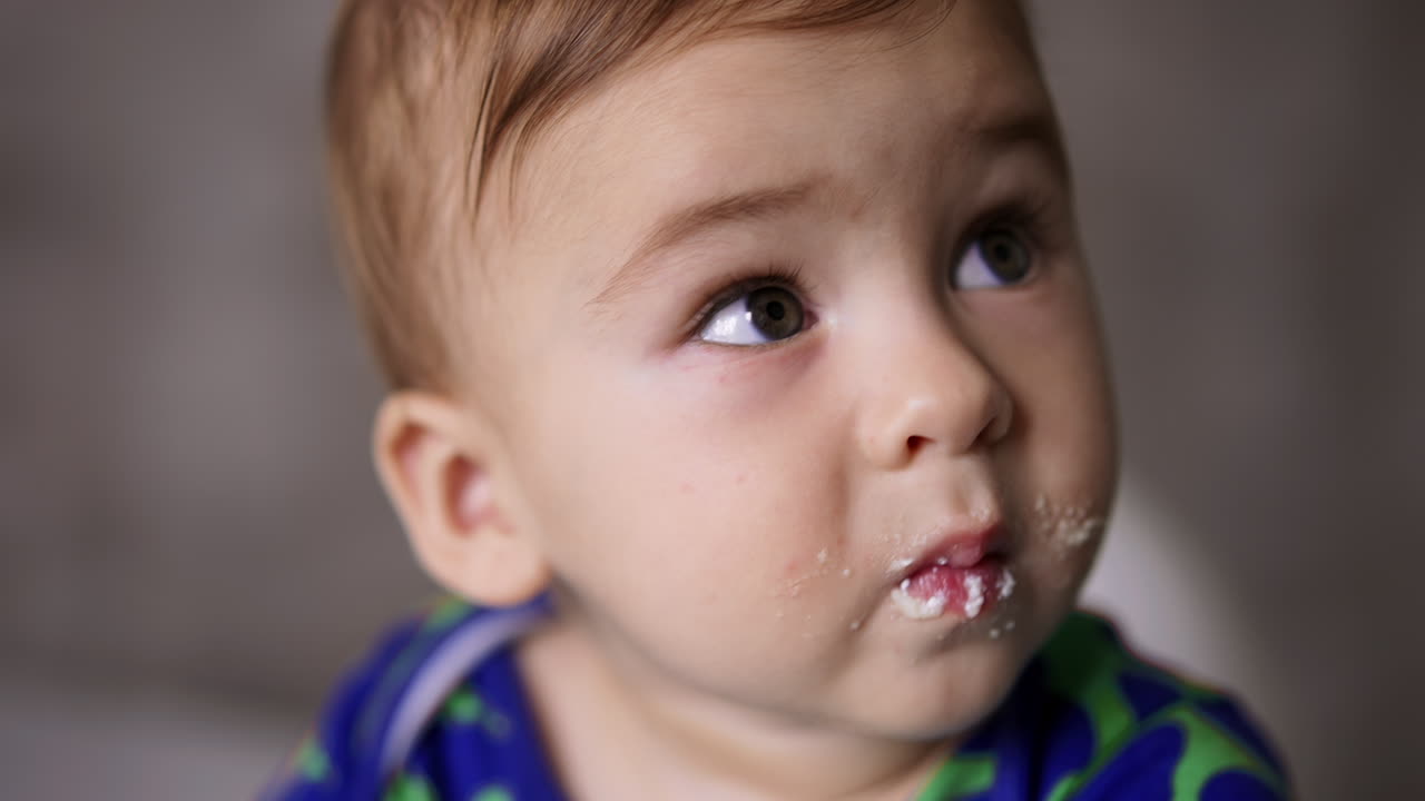 Adorable innocent baby boy with grey eyes and smudged face eating dairy. Feeding little child from spoon. Close up.