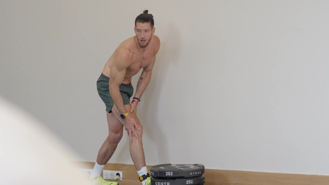 Hombre atlético sin camiseta con moño estirando la pierna y los músculos cuádriceps durante un calentamiento de movilidad contra una pared blanca y limpia en un estudio de gimnasio