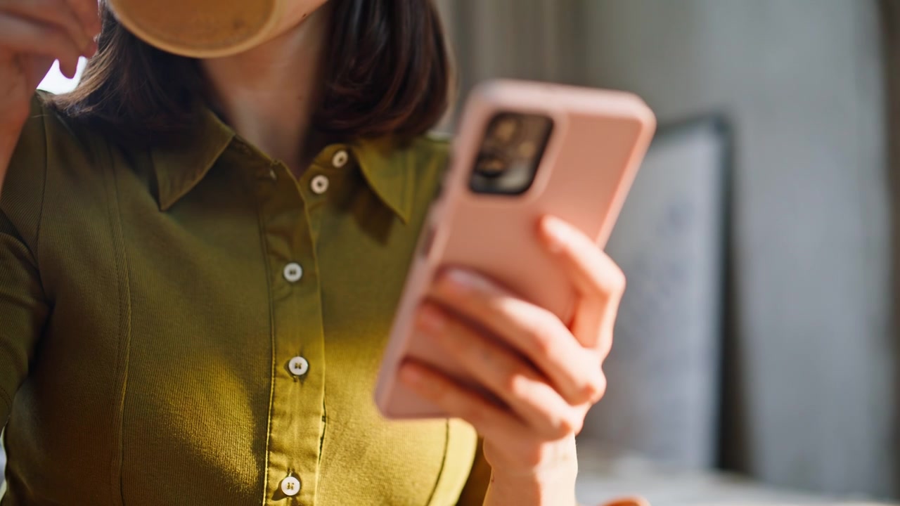 Primo piano: mano che tiene una tazza di tè in un appartamento soleggiato. Bruna che scorre il cellulare riposando.