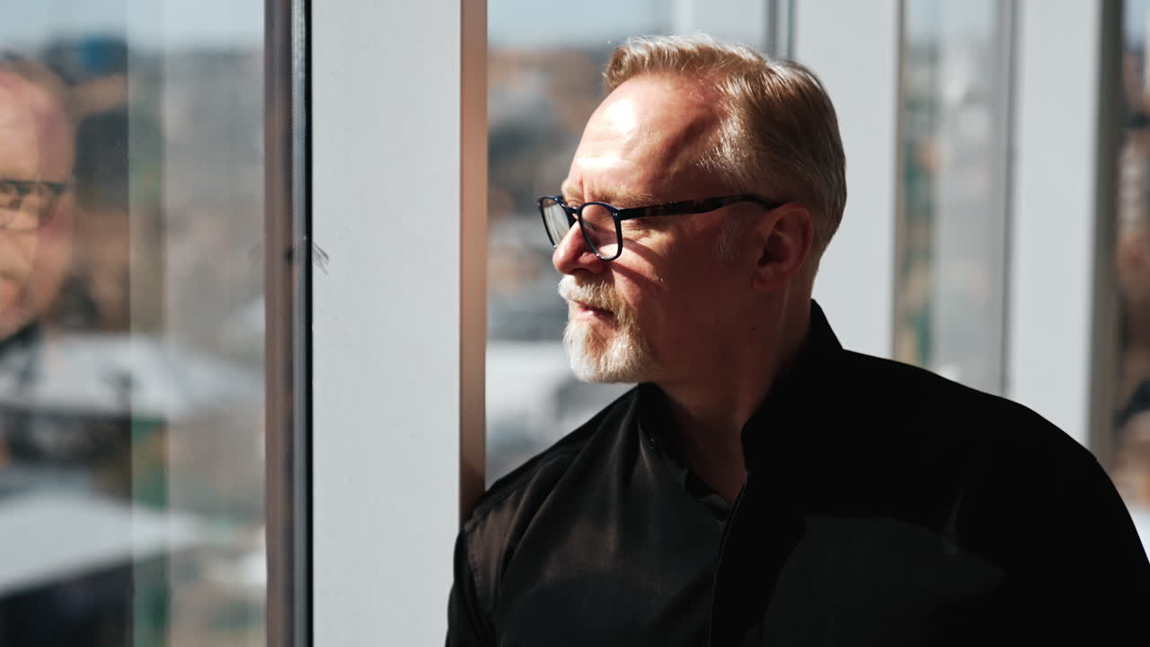 Serious attractive confident mature man in glasses and black shirt in office. Cropped portrait of a businessman standing at window on sunny day looking at cityscape.