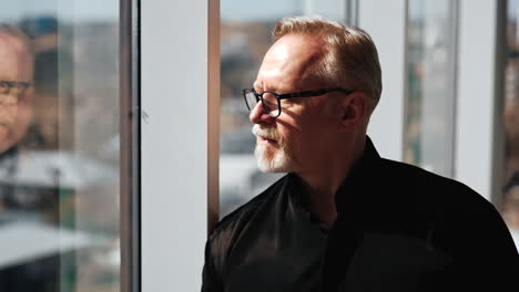 serious attractive confident mature man in glasses and black shirt in office. cropped portrait of a businessman standing at window on sunny day looking at cityscape.