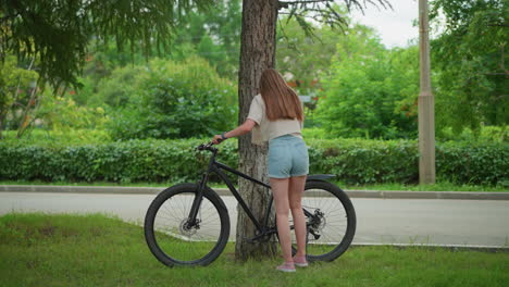 woman in jean shorts approaches bicycle leaning against tree in tranquil park, removes stand, and prepares to move with bike, scene features lush greenery, trees, and a paved path