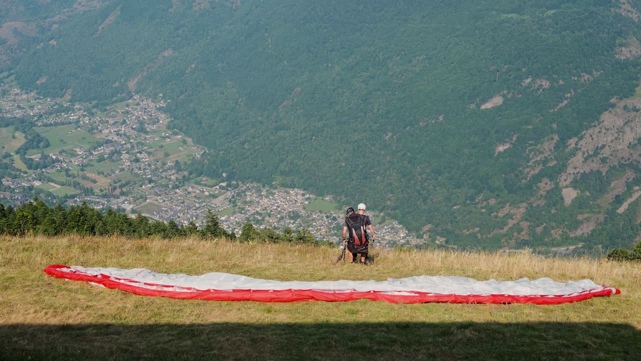 Two paragliders prepare to launch from a grassy hillside with a vast mountain valley below