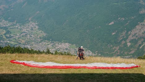 two paragliders prepare to launch from a grassy hillside with a vast mountain valley below
