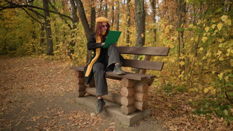 young artist deeply focused on sketching seated on wooden park bench in peaceful autumn park, one leg on bench, one on ground, with bag on floor, surrounded by golden leaves and serene nature