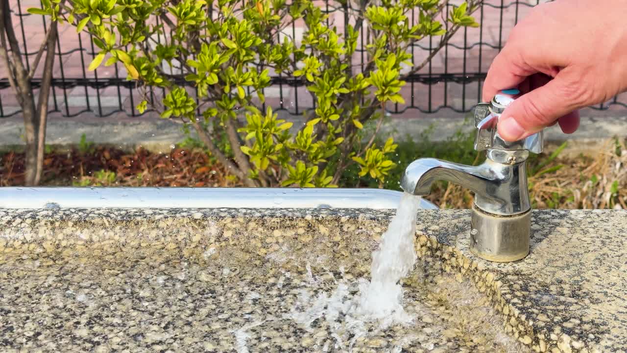 Una mano aziona un rubinetto cromato su un lavabo in pietra in un parco soleggiato.