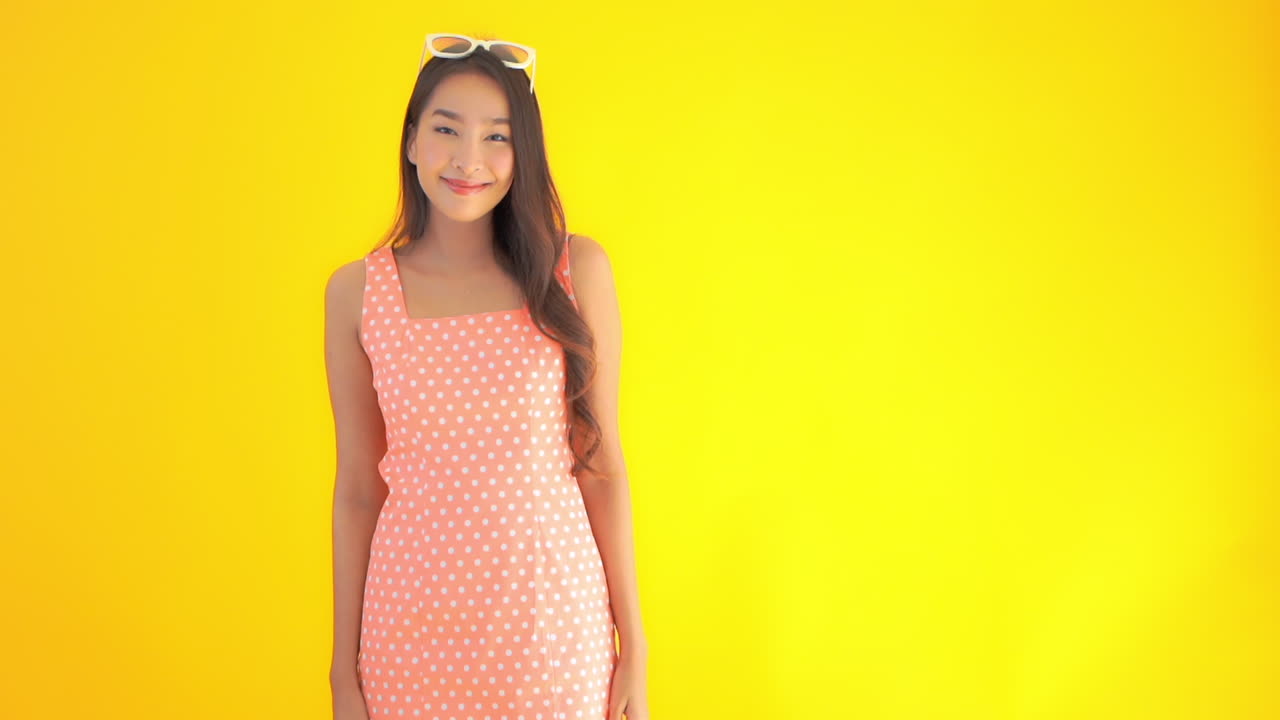 A cute Asian girl dressed in a sundress and sunglasses raises her hands flashing the peace- victory sign