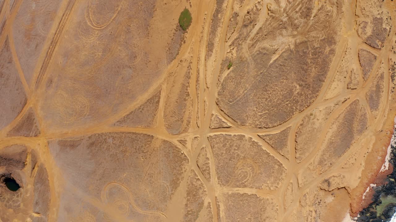 A vertical drone reveals a coastal desert webbed with tire tracks as teal surf slams black rocks, blending human tracery with raw shoreline textures and wind-hardened stillness
