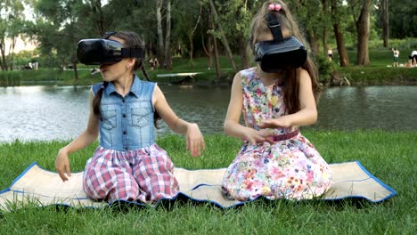 happy kids with virtual reality glasses are sitting in the park on the grass by the river and playing. they swim in a virtual game and laugh.