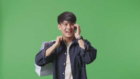 초록색 화면 앞에서 는 동안 전화에 응답하고 휴대 전화로 이야기하는 쇼핑 가방을 가진 아시아 쇼핑 남자