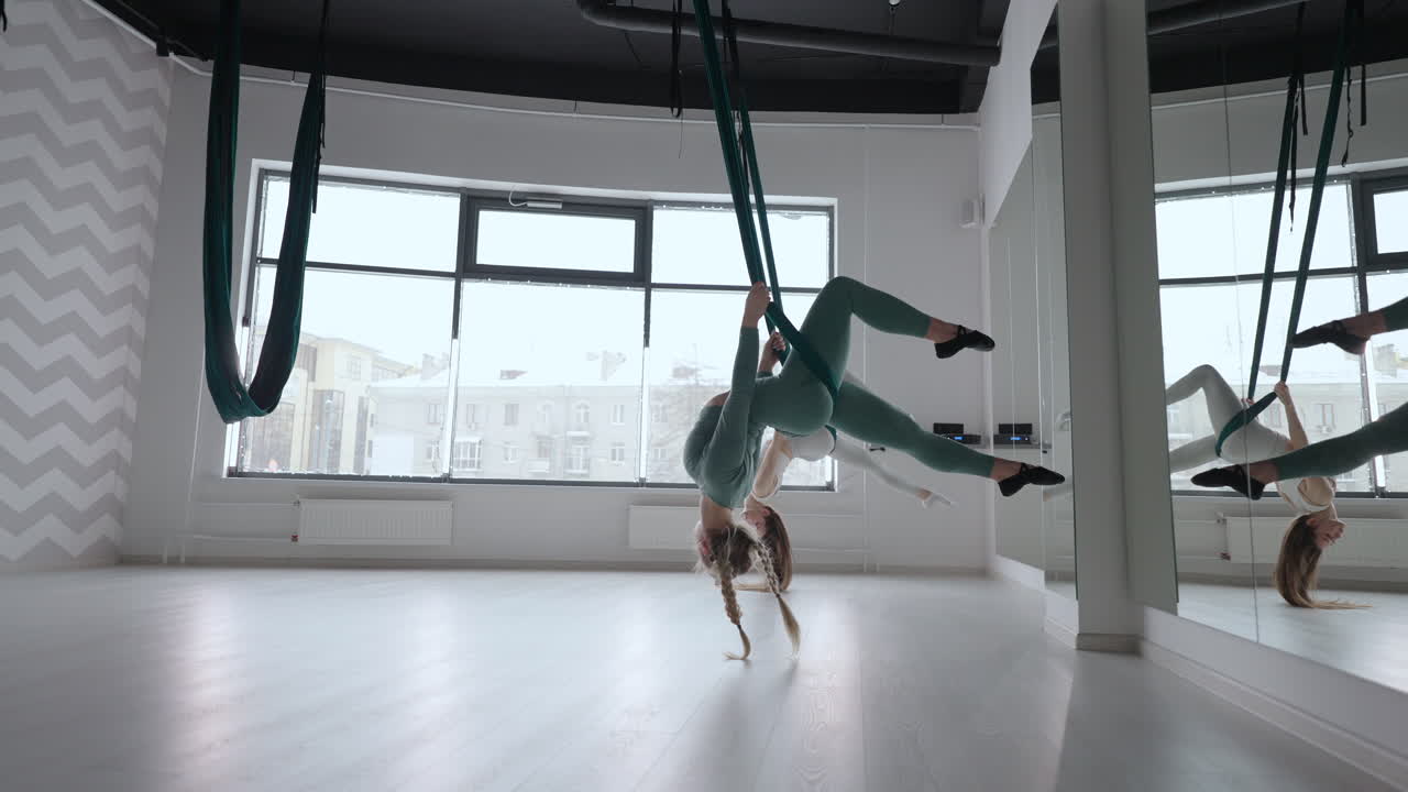Due giovani donne yogi praticano yoga aereo in amache verdi in una palestra. Belle donne si allenano in classe eseguendo aero yoga. Variazione della posa della Piramide Parsvottanasana.
