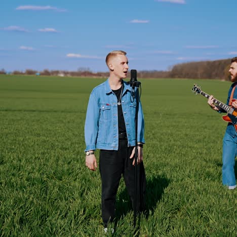 vocalist and his music band among nature. young man singing into a microphone while his friends playing musical instruments on green field in spring