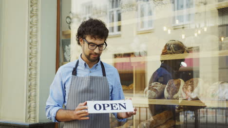 Male-Smiled-Bakery-Vendor-Putting-And-Turning-Signboard-Open-On-The-Glass-Door-Of-The-Shop