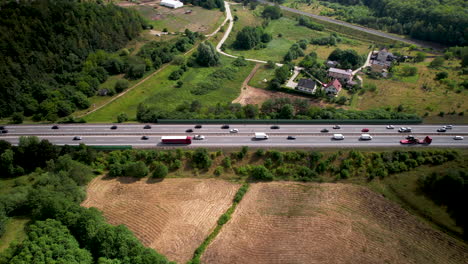 Vista-De-Seguimiento-Aéreo-Del-Tráfico-Fluido-De-Automóviles-Y-Camiones-En-La-Carretera-Rural-De-Varios-Carriles,-Rodeada-De-Campos,-Tierras-Verdes,-árboles-Y-Casas-Suburbanas,-Viajes-Y-Transporte-En-Gdynia,-Polonia