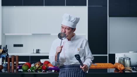 asian man chef holding tongs and ladle and singing while standing in home kitchen