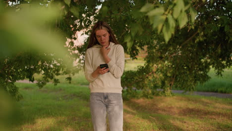 girl in casual clothing stands under tree, focused on phone screen with hand on hood collar, sunlight filters through leaves, casting soft shadows while background greenery sways gently in breeze