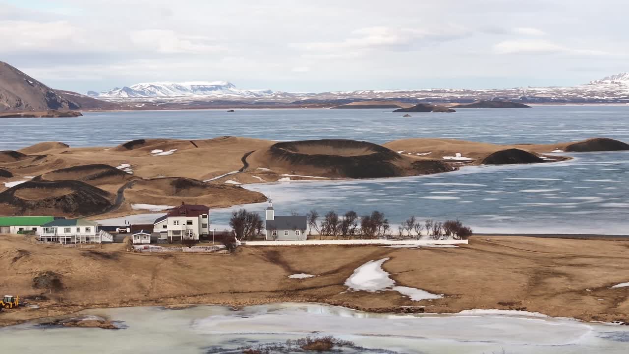 Scenic view of the frozen Lake Mývatn and surrounding volcanic pseudocraters in Skútustaðir, Iceland, with a small village in the foreground.