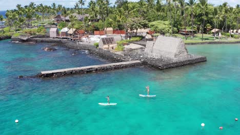 close-up panning aerial shot tracking two stand-up paddleboarders in front of the historic kamakahonu house in kailua-kona on the big island of hawai'i