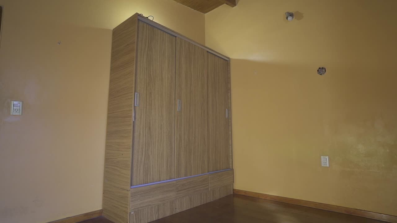 Still camera clip portraying large wooden closet against yellow wall in rental house room, with artificial lighting behind and wooden floor.
