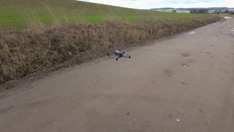 the drone hovers above a muddy path surrounded by tall, dried grass, with fields and a hilly terrain in the distance