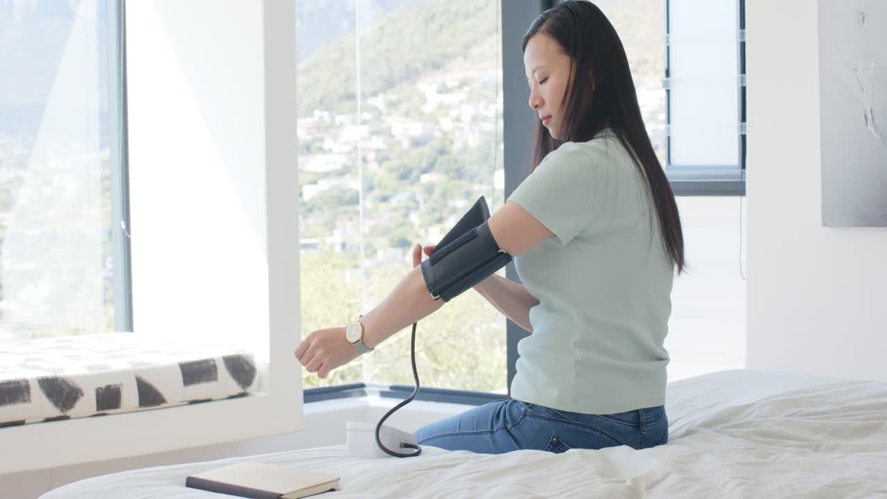 Donna asiatica seduta sul letto vicino alla finestra che controlla l'orologio e prepara un bracciale per la misurazione della pressione.