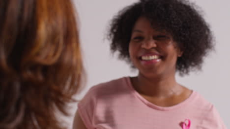 Close-Up-Studio-Portrait-Of-Two-Smiling-Mature-Women-Wearing-Pink-Breast-Cancer-Awareness-Ribbons-Hugging-Against-White-Background