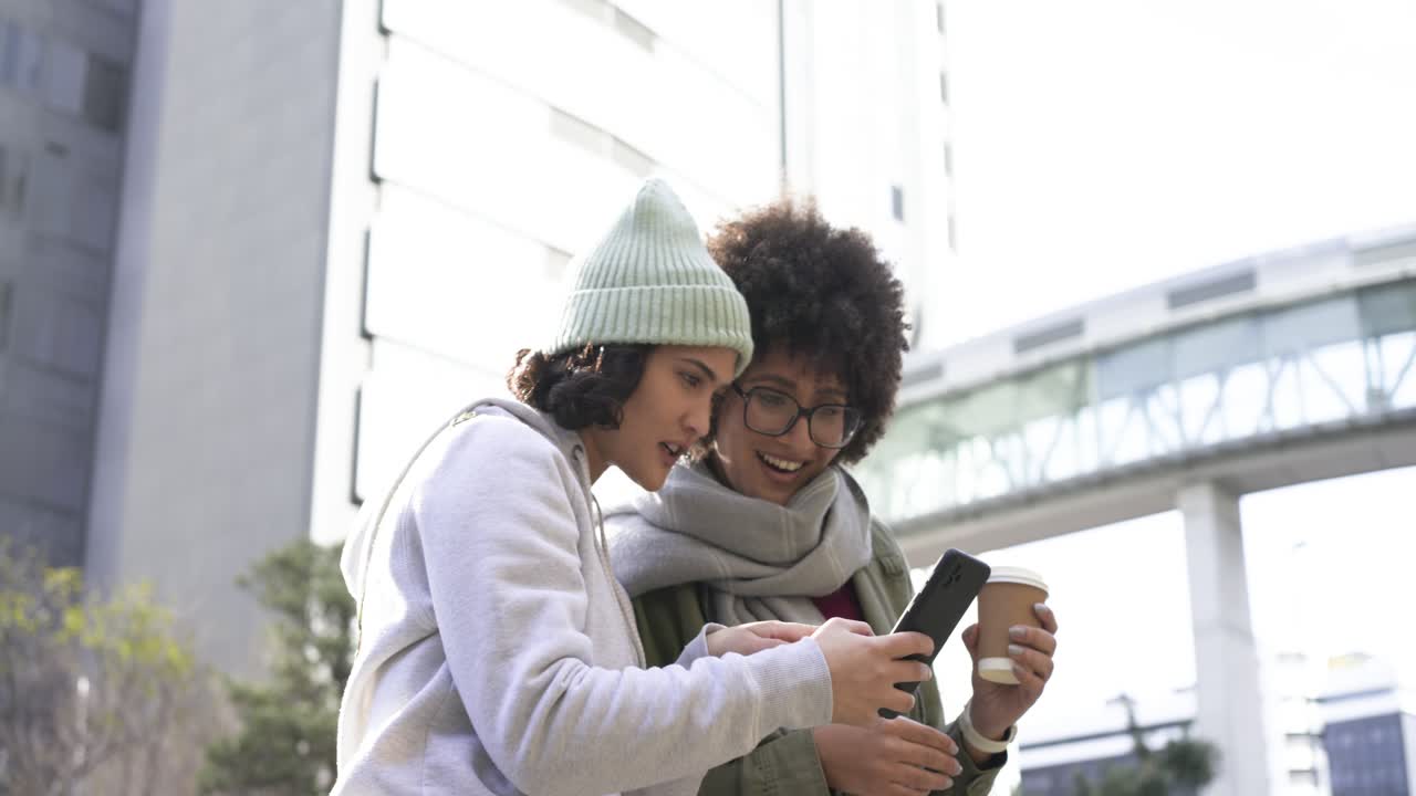 Female friends at plaza seeking directions on smartphone, pointing at skybridge, holding coffee