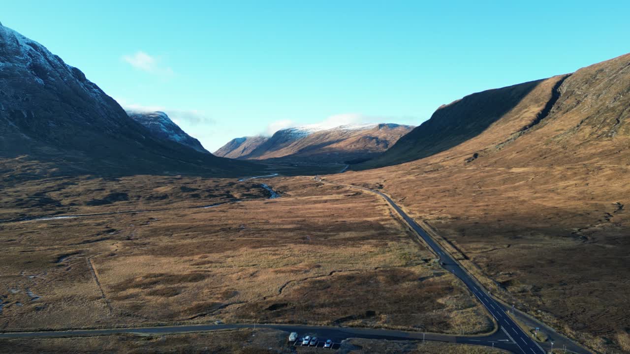 Valle de Glencoe con carretera serpenteante, serenas Tierras Altas escocesas, luz del día, vista aérea.