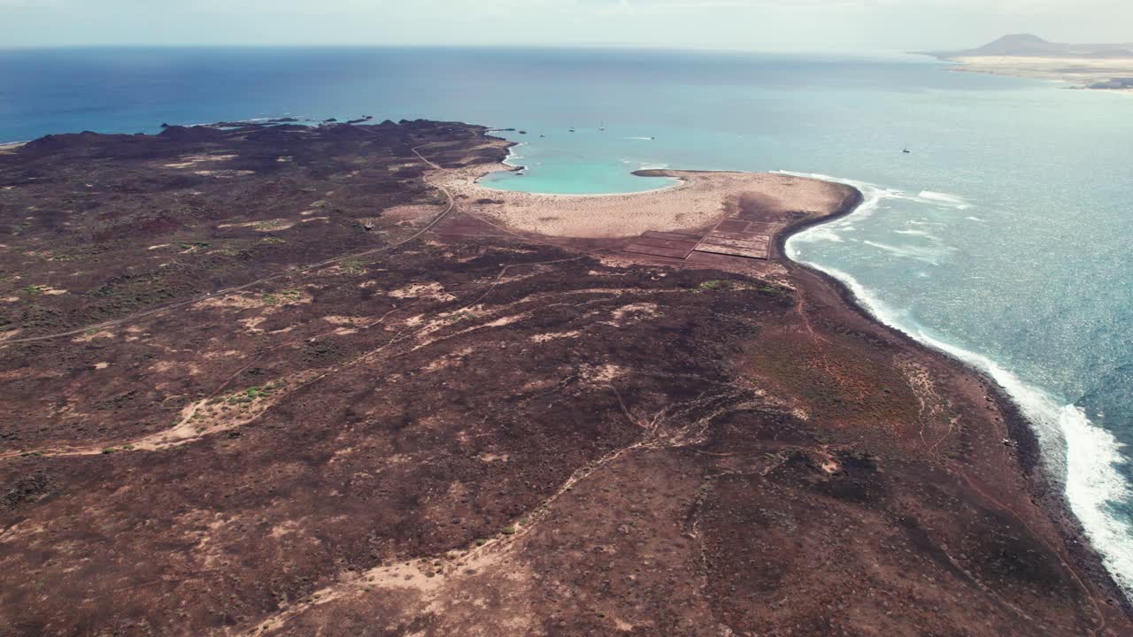 Drone aerial view of Playa De La Concha De Lobos, Lobos Island, capturing rugged volcanic terrain meeting turquoise waters and a pristine curved beach under a clear sky.
