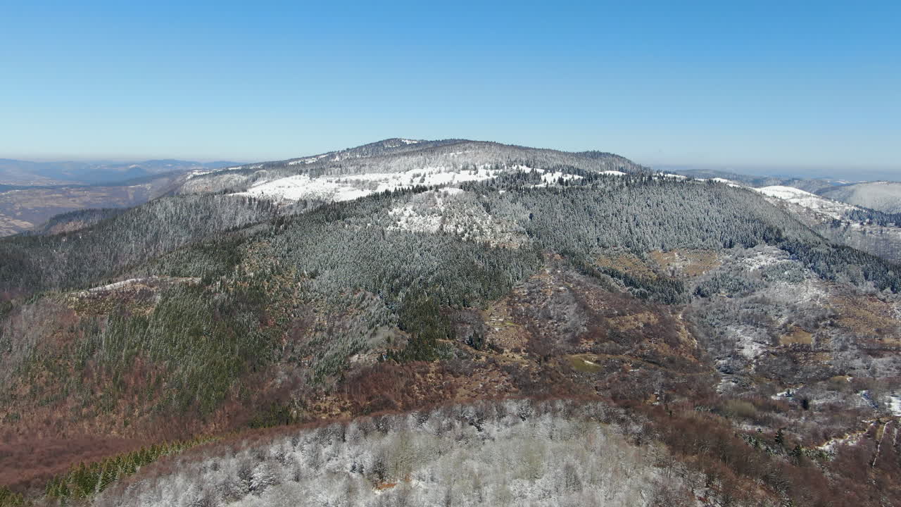 View of a mountain range with areas covered in snow and trees The sky is blue The terrain varies in color