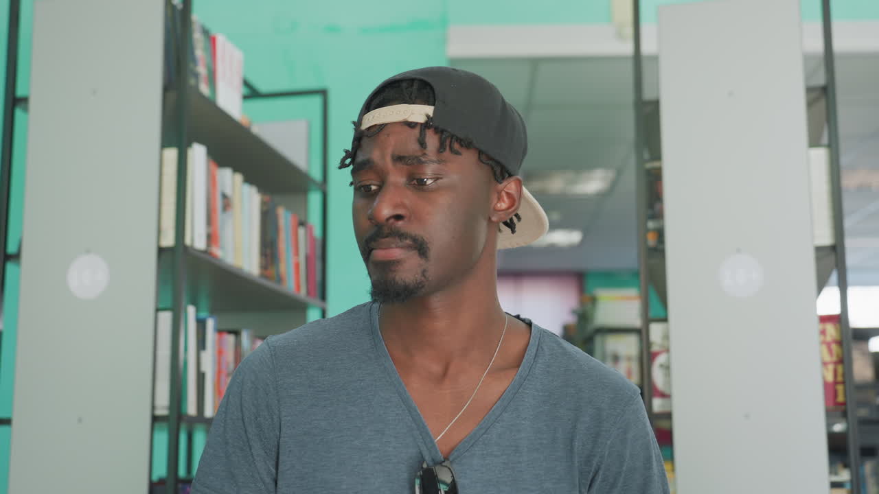 Writer in casual outfit with cap backwards and sunglasses on shirt sits in library aisle reading book surrounded by tall bookshelves filled with colorful books in quiet well lit indoor space