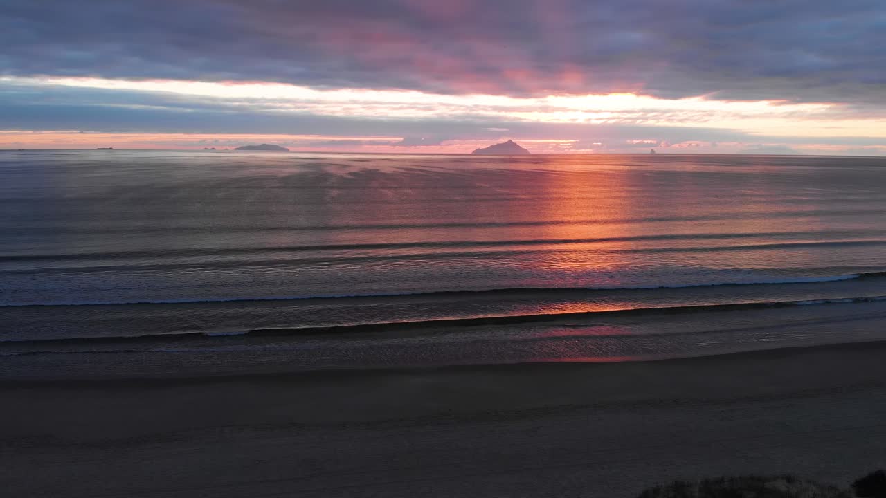 volando hacia atrás sobre una playa y un océano en calma, siempre más alto, con islas y un amanecer nublado