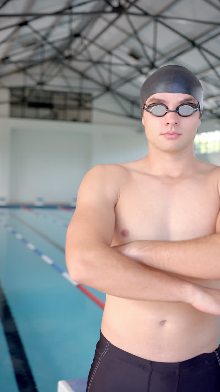 Vertical video: Swimmer in swim cap and goggles standing confidently by indoor pool