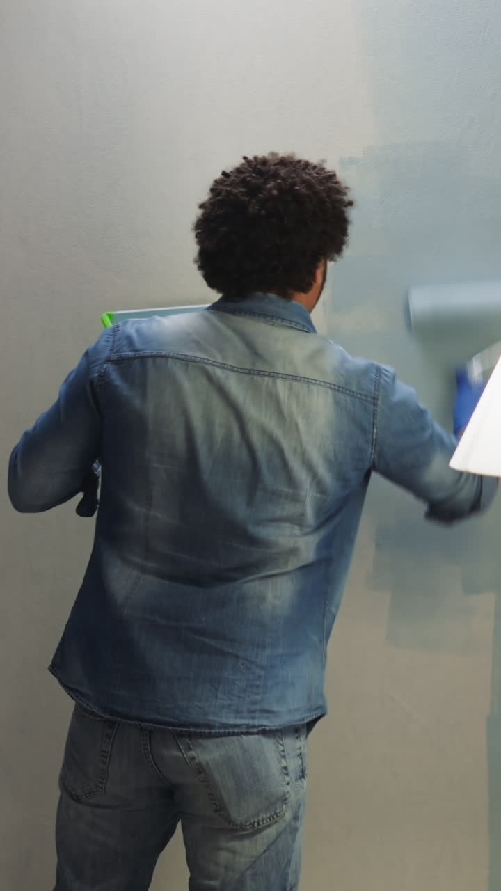 Black man in denim shirt paints wall with blue color in living room backside view. Young African-American worker in jeans renovates apartment interior