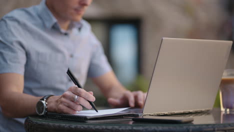 un hombre sentado en un café de la calle trabaja de forma remota escribiendo un bolígrafo en un cuaderno. trabaja en la computadora portátil. un hombre trabaja de forma remota en una computadora portátil