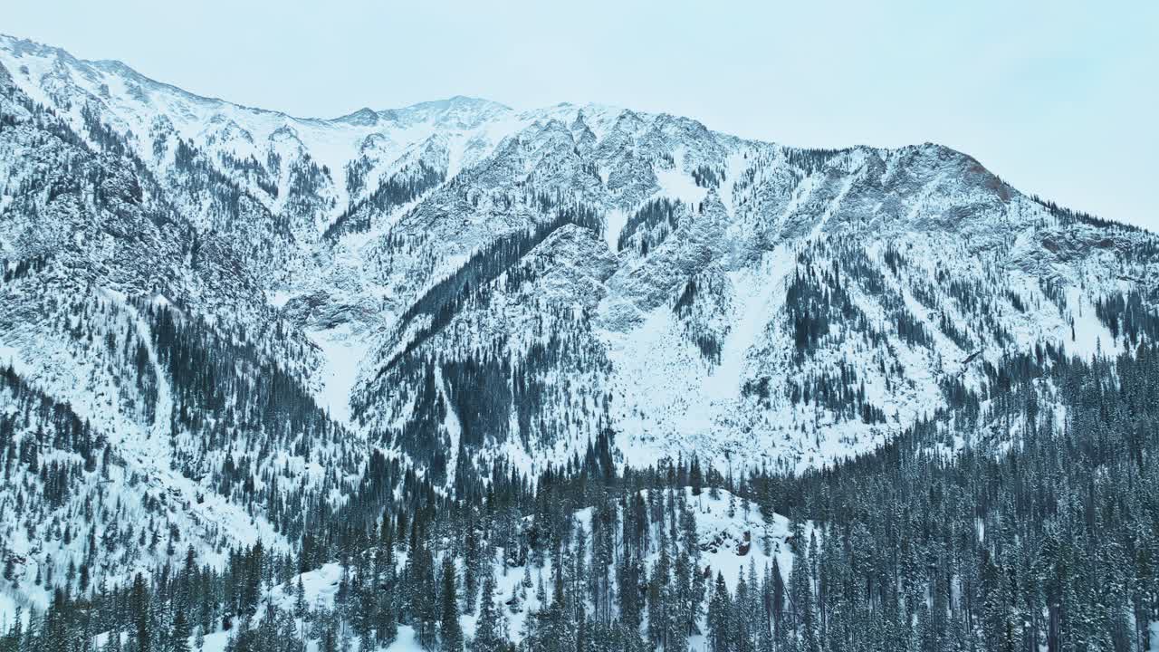 Aerial sweeping over snow covered mountain ridges with dense evergreen forests, winter scenery in rugged terrain, pullback reveals hillside