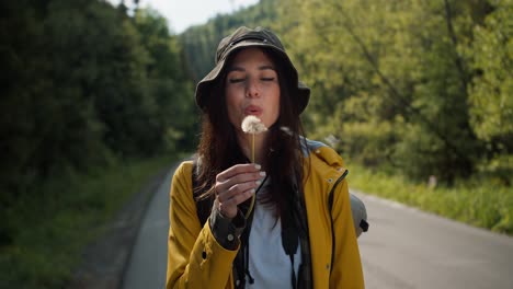 feliz chica morena turista en ropa especial para hacer senderismo en una chaqueta amarilla sopla en un diente de león que está volando contra el fondo del bosque