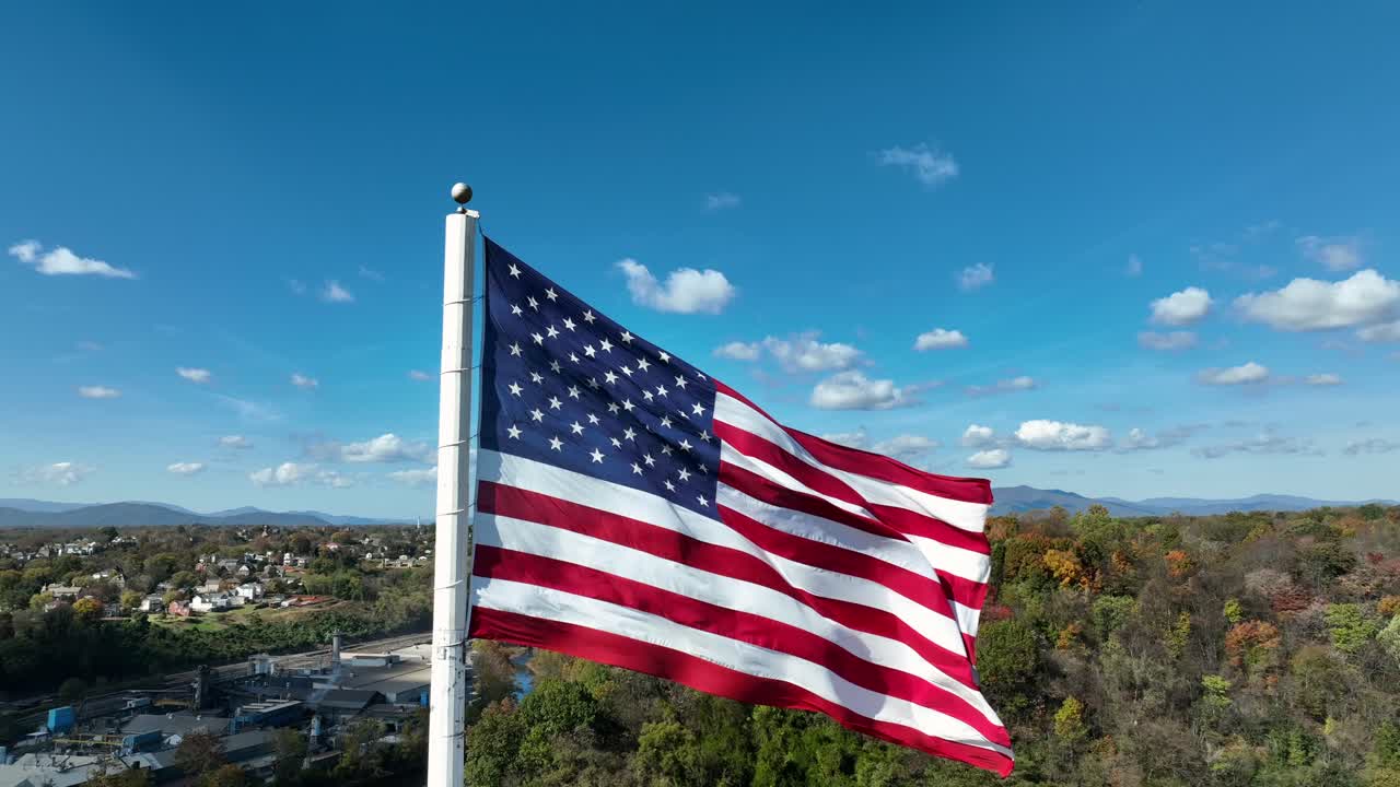 Proud American flag waving on flagpole over colorful trees in autumn season. Blue sky in suburb district of American town. Aerial close up orbit shot.