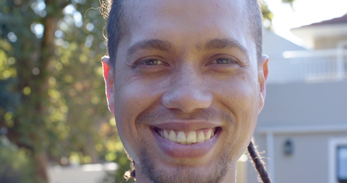 Homem afro-americano parado em frente à casa sorrindo para a câmera, revelando os dentes sob luz de contorno.