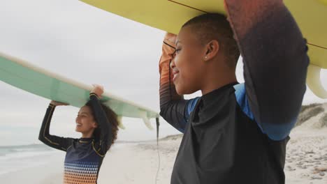 happy african american female friends on the beach holding surfboards