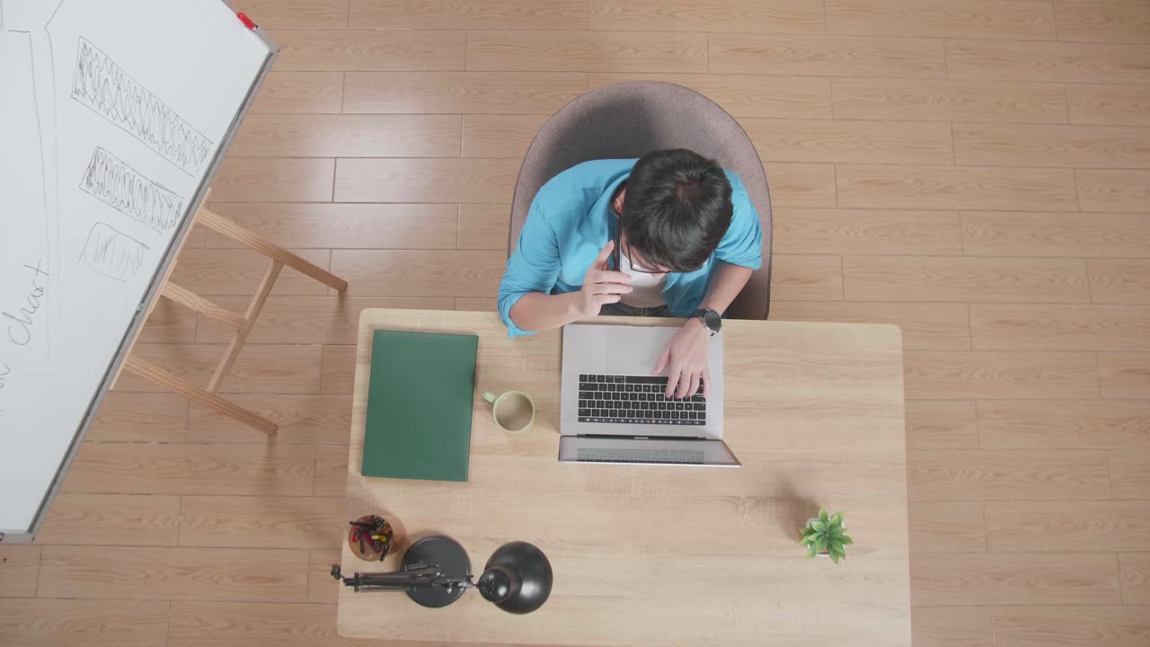 위쪽 뷰  아웃  한 남자가 집에 있는 노트북에서 타입하는 화이트보드 에 있는 나무 책상 에 앉아 스마트폰으로 이야기하고 있다.