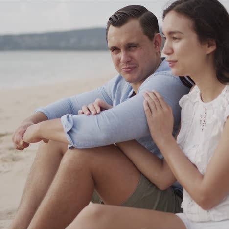 Couple-on-vacation-sitting-on-sand-of-seashore-and-smiling