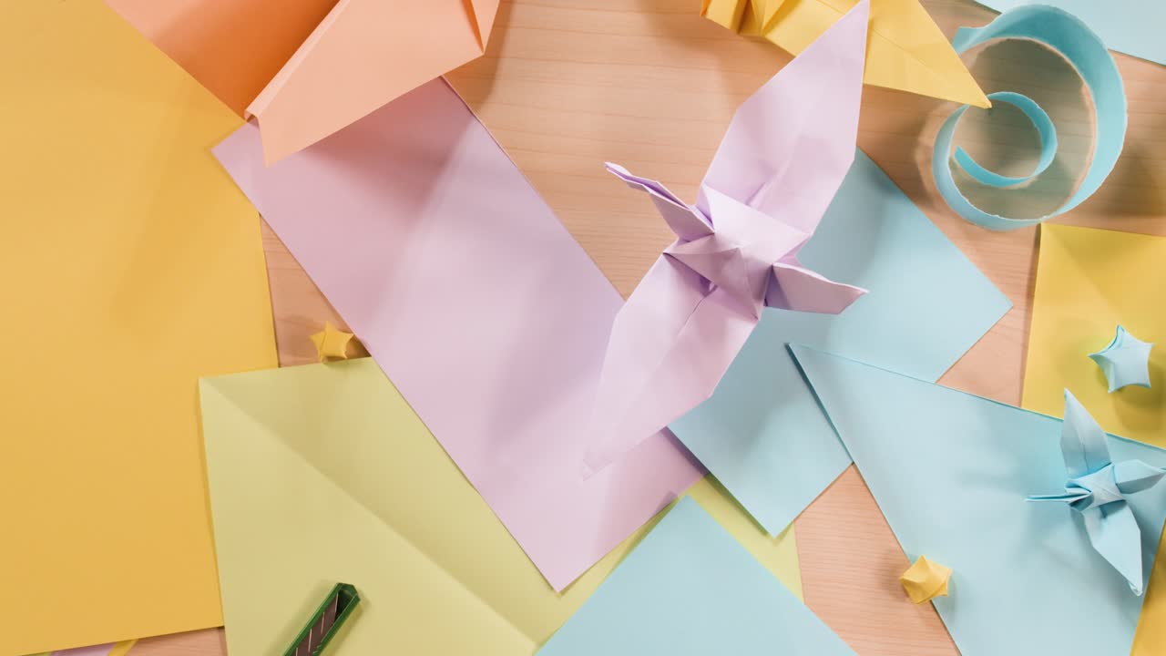 Vista dall'alto di mani che sistemano creazioni origami color pastello e strumenti per l'artigianato su una superficie di legno