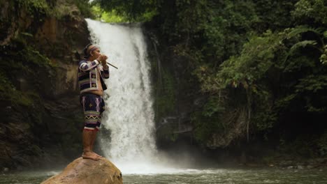 indígena tocando el instrumento tribal kubing, detrás hay una cascada mientras está de pie