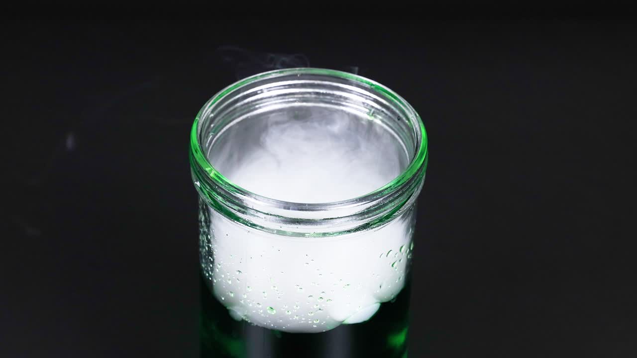 건조한 얼음이 유리 컵에 어지는 장면, 어두운 배경에 회전하는 증기 패턴을 만니다.